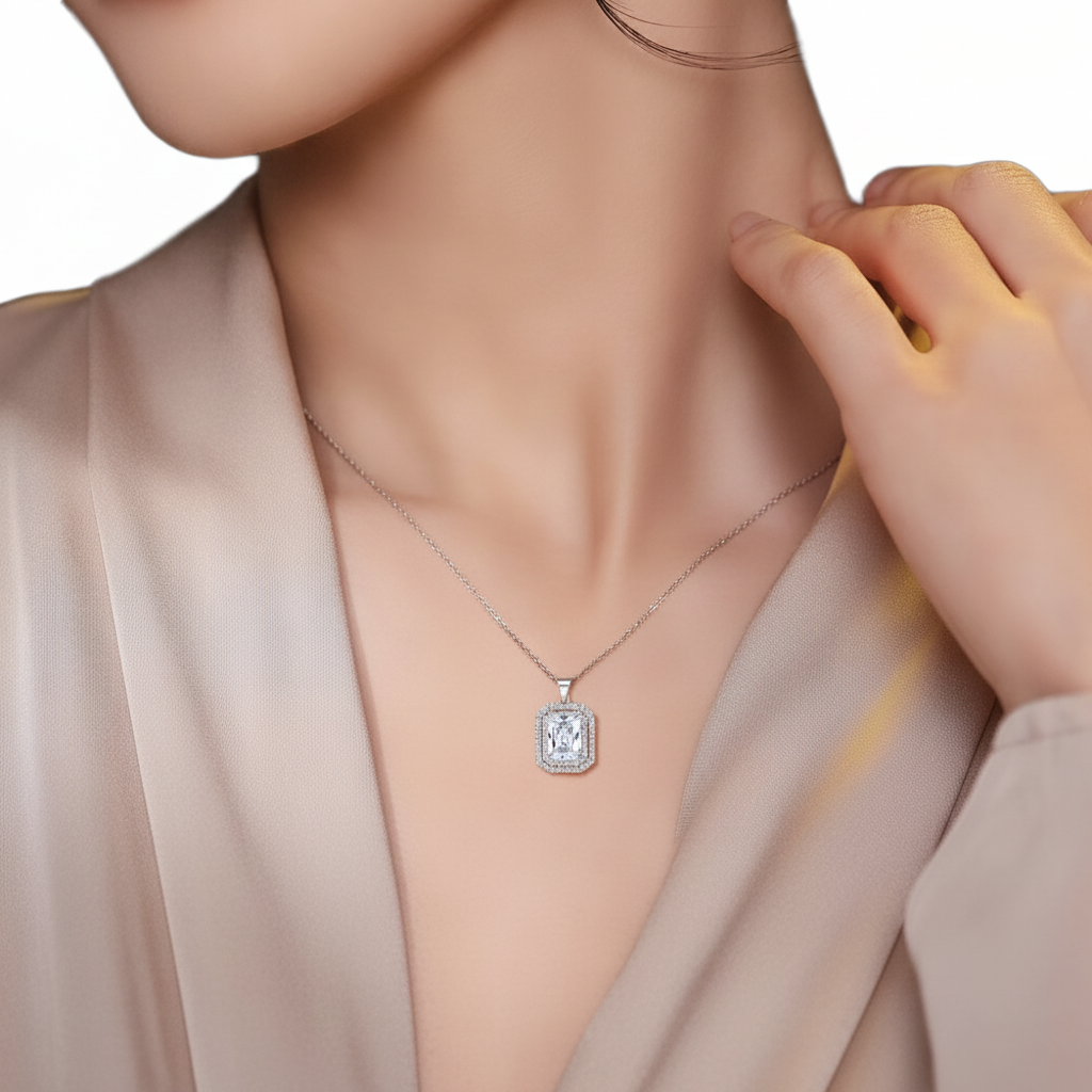 A close-up of a model wearing a silver-toned rectangular pendant necklace with a clear central gemstone, styled with a beige satin blouse against a white background.