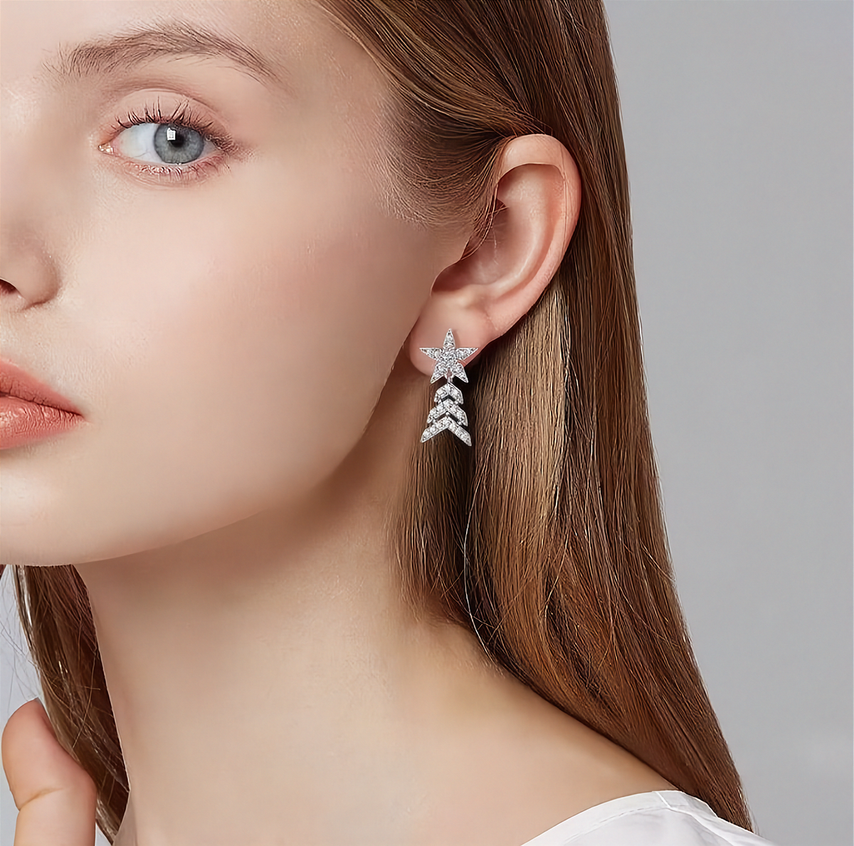 A close-up of a woman's ear wearing a silver dangling earring with a star-shaped top and three chevron drops, set with small clear stones against a soft gray background.