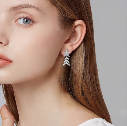 A close-up of a woman's ear wearing a silver dangling earring with a star-shaped top and three chevron drops, set with small clear stones against a soft gray background.