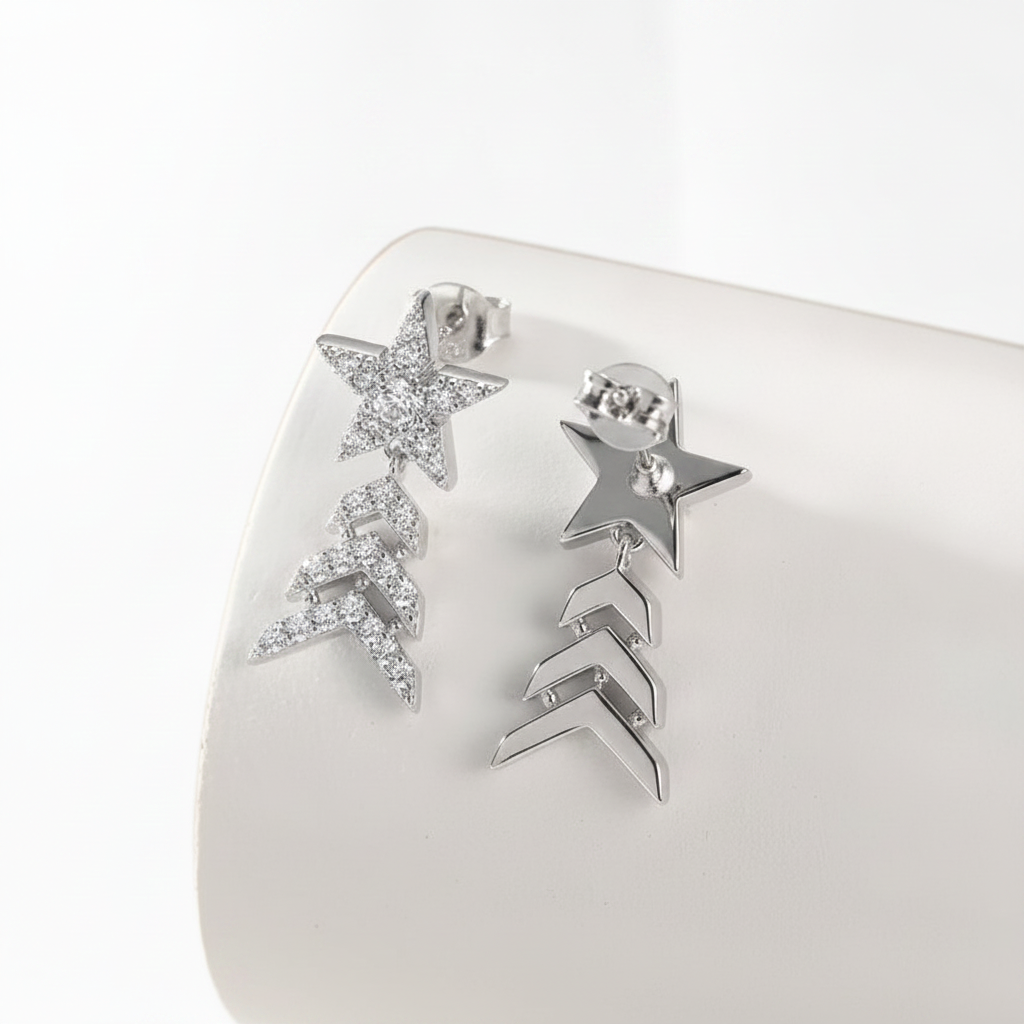 A pair of silver-toned dangle earrings displayed on a white curved jewelry stand. One earring features a star-shaped cluster covered in small clear rhinestones, and the other features a star above three chevron links.