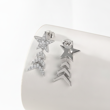 A pair of silver-toned dangle earrings displayed on a white curved jewelry stand. One earring features a star-shaped cluster covered in small clear rhinestones, and the other features a star above three chevron links.